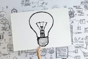 A hand holds up a giant lightbulb sketch, in front of a second board covered in brainstorming sketches and ideas.